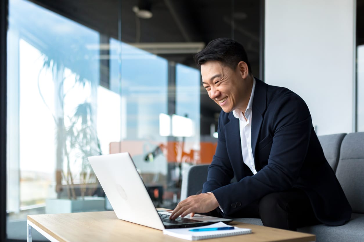 Image of eastern Asian man smiling at a computer in a glass window office