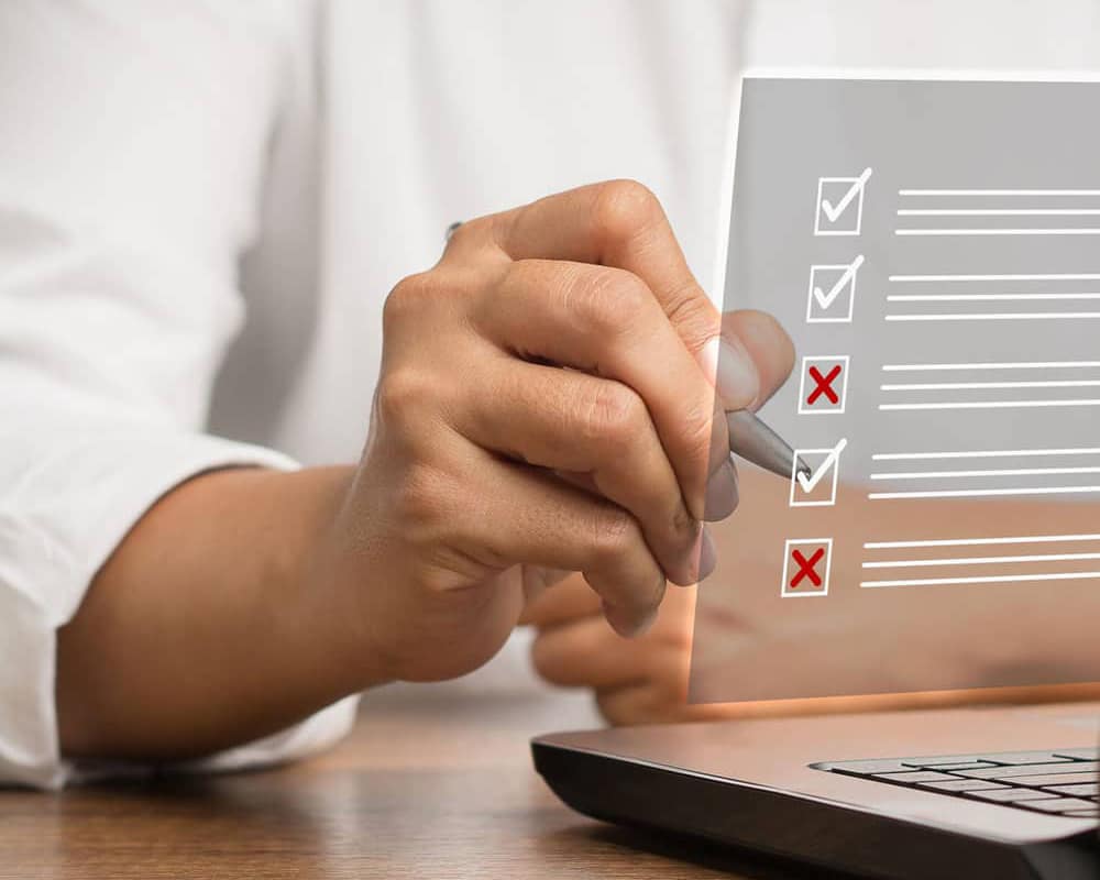 image of a man's hand holding a pen at a computer with a digital overlay of. checklist.