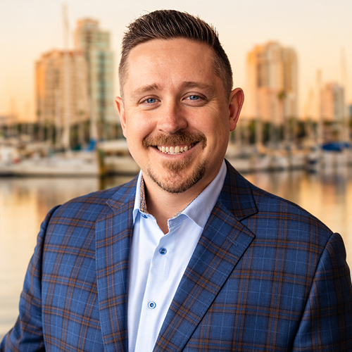 Smiling man in a blue plaid blazer and light blue shirt standing by a marina at sunset.