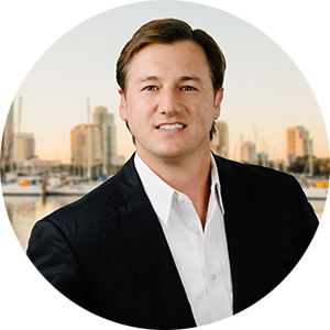 Professional headshot of a man in a dark blazer and white shirt, standing outdoors by a marina with city buildings behind him, smiling.