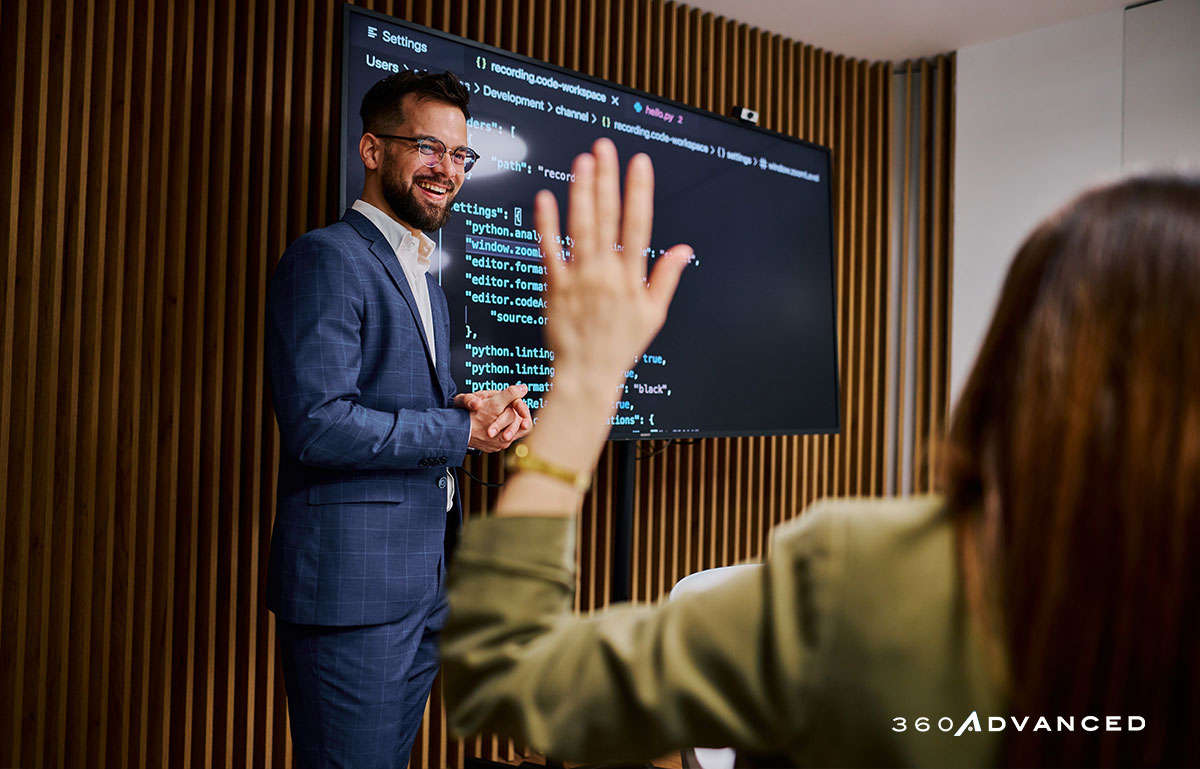 Businessman explaining code on a monitor to his colleagues during a meeting. Woman's hand is raised for a questions in foreground.
