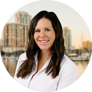 Portrait of a smiling woman with long dark hair in a white blouse, with a waterfront cityscape and buildings in the background.