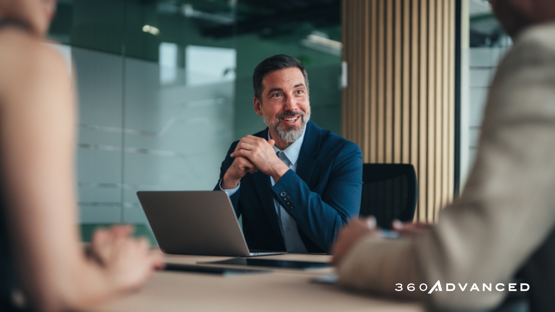 Office professionals in meeting; man sitting at desk in front of laptop talking with two people outside photo frame